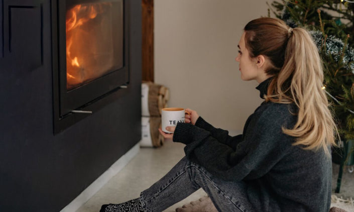 a girl is holding a cup of tea and sit beside the fireplace